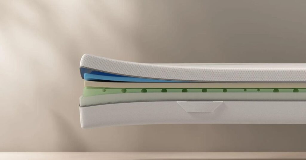 Un materasso in memory foam è posizionato su una base di legno chiaro. Accanto, si vedono cuscini bianchi e una coperta piegata. L'ambiente è luminoso e accogliente.