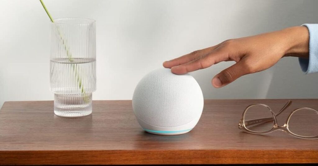 Un Amazon Echo si trova su un tavolo di legno in un soggiorno luminoso, circondato da piante verdi e un divano grigio. La luce naturale entra dalle finestre, creando un'atmosfera accogliente.