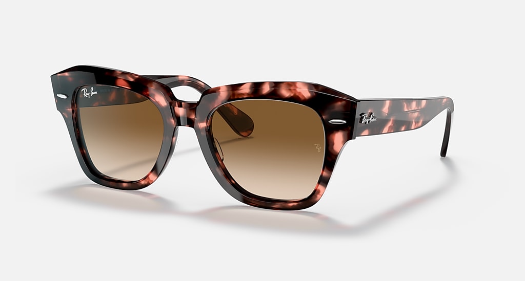 Un parco soleggiato con persone che si rilassano su coperte e sdraio. Alcuni bambini giocano con un pallone, mentre altri adulti chiacchierano. Sullo sfondo, alberi verdi e un cielo blu.