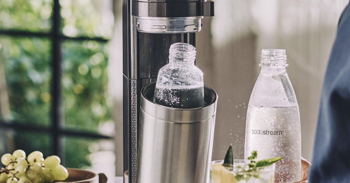 Una persona utilizza una SodaStream per preparare acqua frizzante in una cucina moderna, circondata da bottiglie di sciroppo e bicchieri. L'ambiente è luminoso e accogliente.