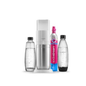 Un elegante gasatore d'acqua SodaStream bianco è posizionato su un piano di lavoro in cucina, circondato da bottiglie di acqua e un bicchiere. La luce naturale illumina l'ambiente.